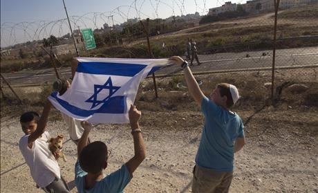 Jewish settler boys hold up an Israeli flag next to the fence of the West Bank settlement of Beit El as Palestinians walk past